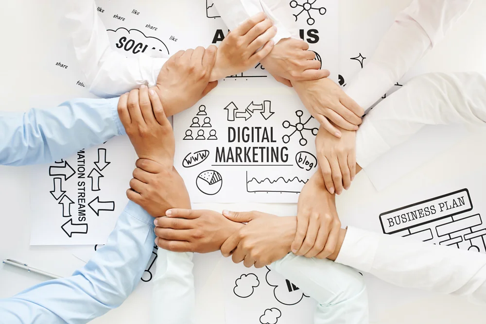 A group of people join hands in a circle over papers with digital marketing and business plan diagrams.