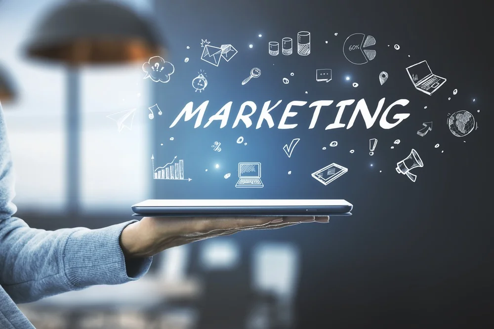 A hand holding a tablet with the word "MARKETING" and various marketing-related icons, such as a lightbulb, graph, and megaphone, floating above it.