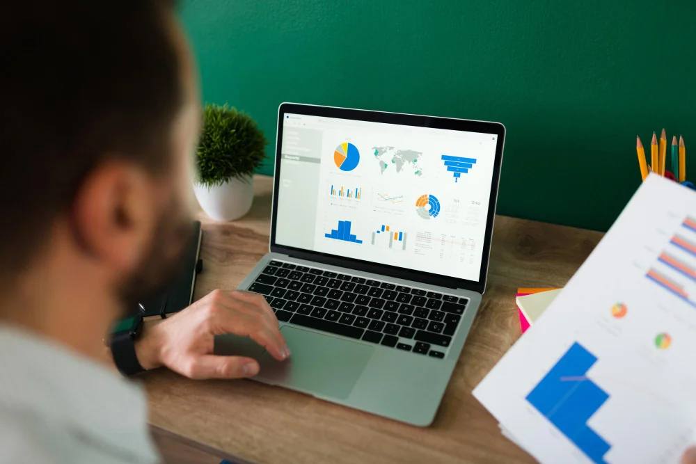 A person reviews charts and graphs on a laptop screen while holding a document with similar data at a desk.