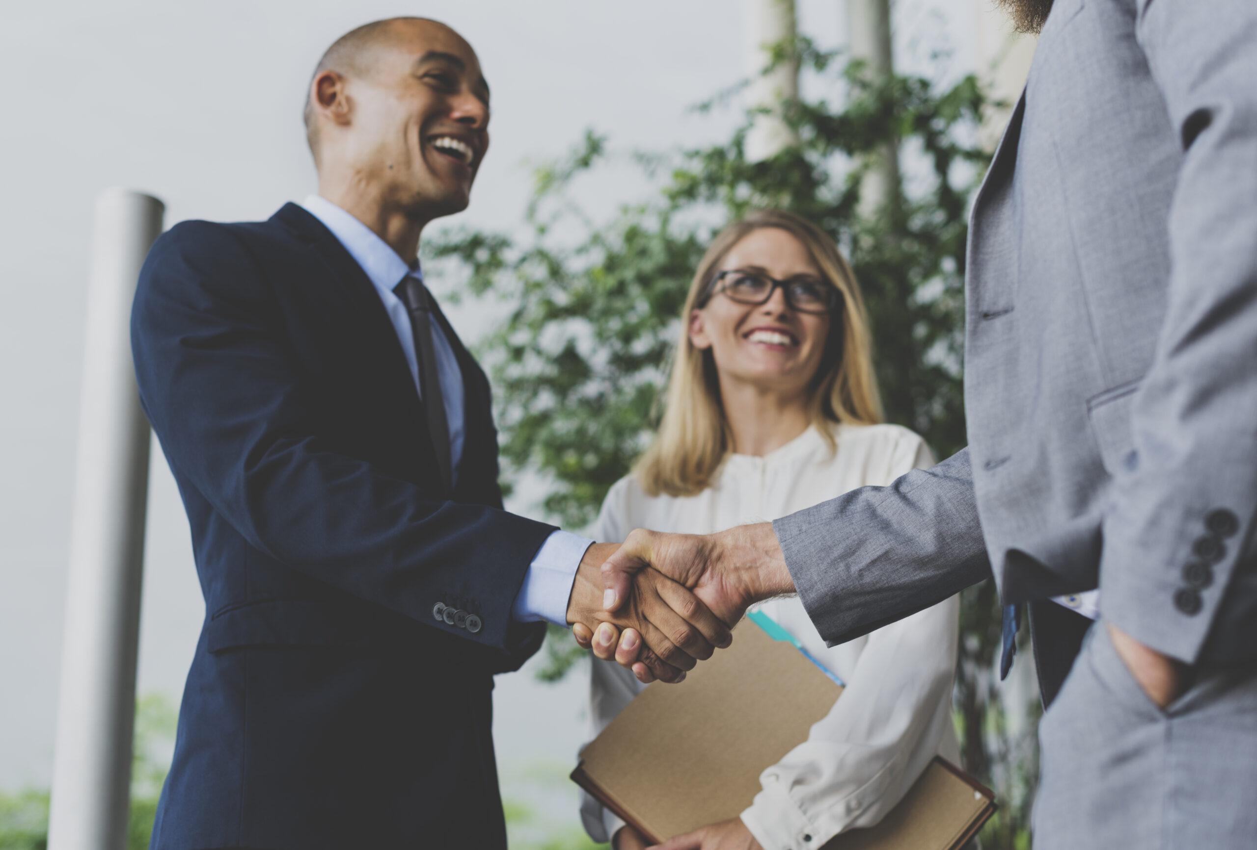 Two men in suits shake hands while a woman holding a folder stands nearby smiling, all in a professional outdoor setting.
