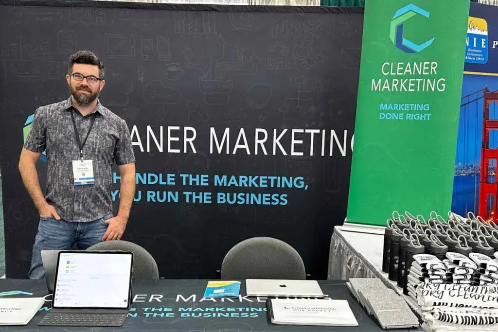 A man stands at a trade show booth for Cleaner Marketing, with promotional materials and company-branded items displayed on the table.