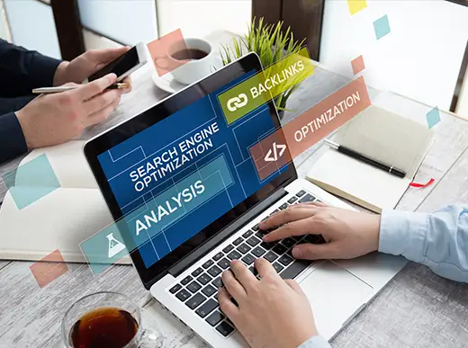 Two people work at a desk with a laptop displaying "Search Engine Optimization" and related terms like backlinks, analysis, and optimization. Notebooks, coffee, and a plant are visible.