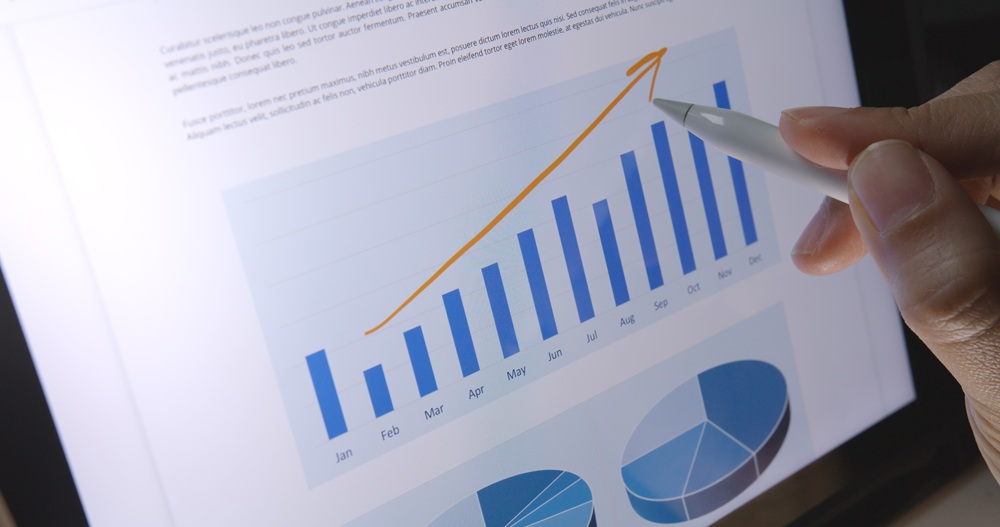 A hand uses a stylus to point at a tablet screen displaying a bar chart with rising values, an upward-trending line, and pie charts below.