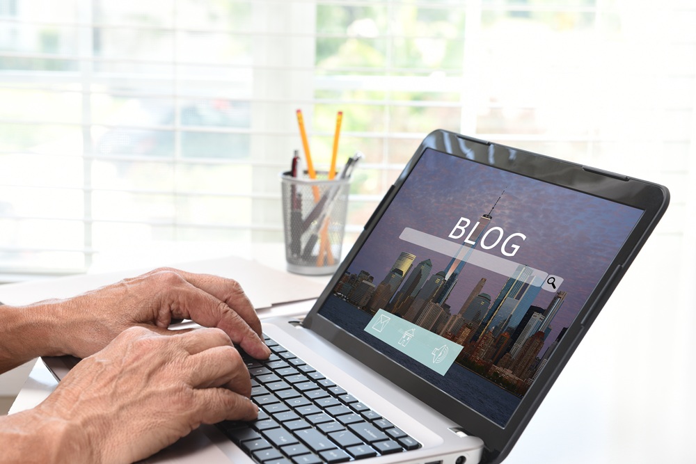 Person typing on a laptop showing a "BLOG" search page with a cityscape image in the background; desk with office supplies and window blinds behind.