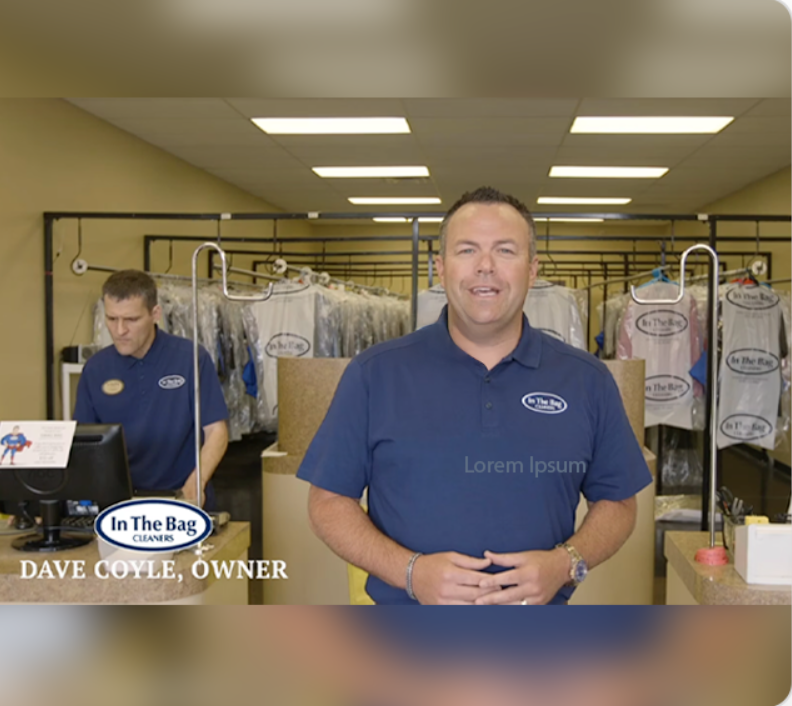Two men in blue "In The Bag Cleaners" shirts stand inside a dry cleaning shop; one is at a counter, the other works at a computer. Clothing is visible hanging in the background.