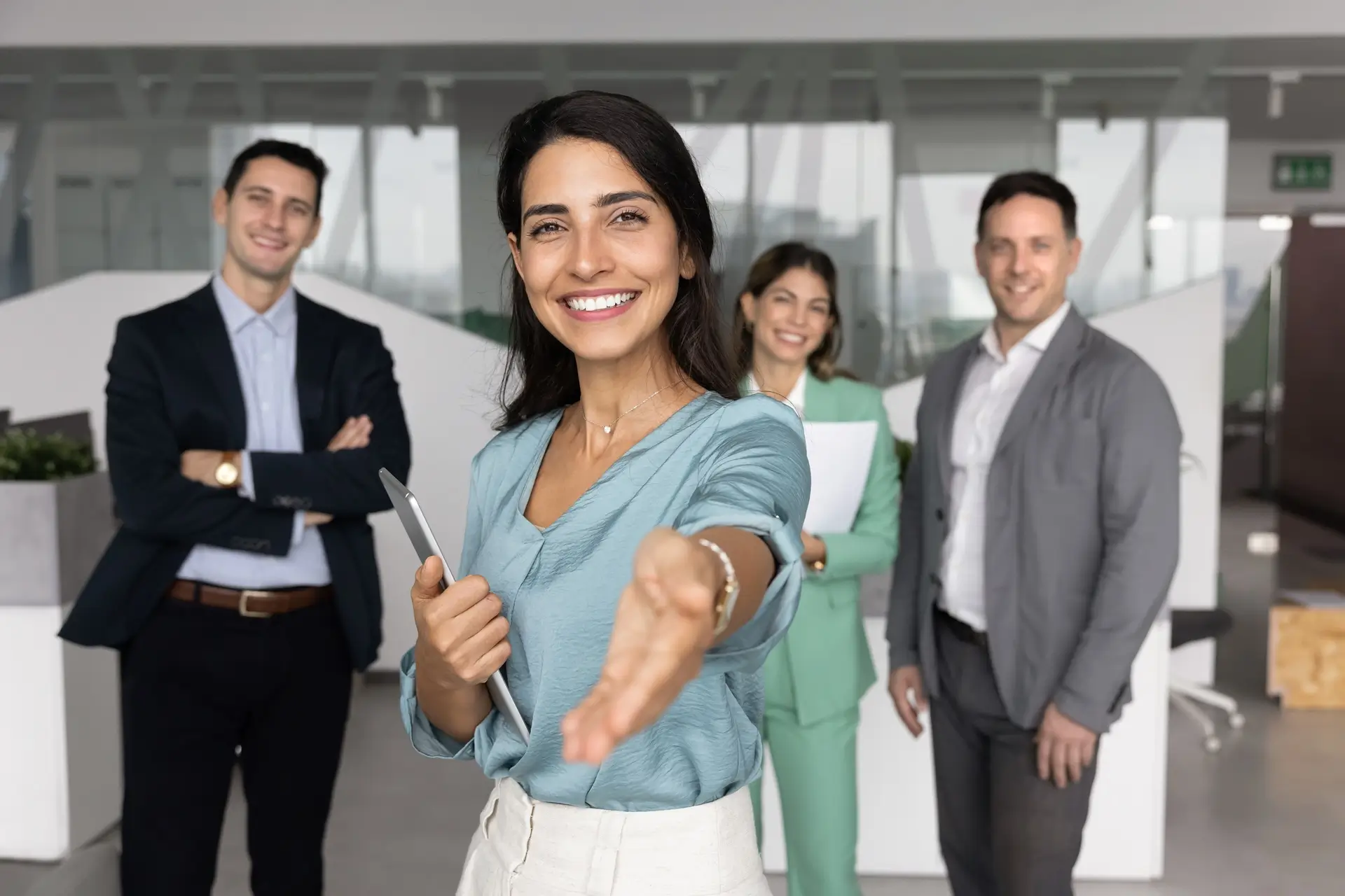 A woman smiles and extends her hand for a handshake, standing with three colleagues in business attire in a modern office setting.